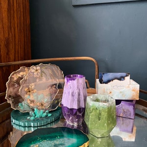 May include: A set of four green, purple, and white resin coasters with gold accents. The coasters are shaped like agate slices and have a geometric design. One coaster features a pressed flower design.