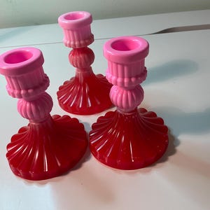May include: Three decorative candle holders. The bases are a vibrant red, with a fluted design. The upper sections are a soft pink, with a rounded, ribbed design. The candle holders are made of a glossy material and are arranged on a white surface.