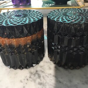 Könnte beinhalten: Zwei schwarze und goldene Dekorationsbehälter mit einem türkisfarbenen und glitzernden Deckel. Die Behälter haben ein geometrisches Design und sind aus Harz.