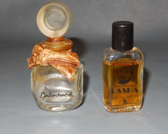 Puede incluir: Dos frascos de perfume vintage. Uno es un frasco pequeño, transparente y cuadrado con la palabra "Cabochard" en cursiva. El otro es un frasco rectangular con tapa negra y la palabra "PASHA" en letras doradas. Ambos frascos están sobre una superficie gris.
