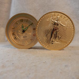 Puede incluir: Un reloj Bulova dorado con números romanos y una moneda de oro con una mujer sosteniendo una antorcha y una rama de olivo. El reloj tiene una esfera redonda y manecillas que indican la hora. La moneda está al lado del reloj.