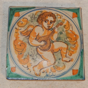 May include: Square ceramic tile featuring a cherub playing a lute. The cherub is depicted in shades of orange and brown, set against a backdrop of green and blue. The tile has a green border with orange accents and a signature in the bottom right corner.