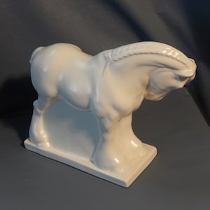 Puede incluir: Una figurita de caballo de cerámica blanca con una crin detallada y un acabado brillante. El caballo está de pie sobre una base rectangular.