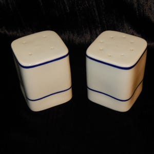 May include: Two white ceramic salt and pepper shakers with a square design. Each shaker has a blue line around the top and bottom, and small holes on the top. The shakers are on a black background.
