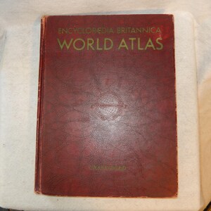 May include: A vintage World Atlas book with a textured, burgundy cover. The title "ENCYCLOPEDIA BRITANNICA WORLD ATLAS" is printed in green. The book appears well-worn, suggesting it is an antique or a used item.