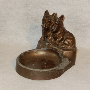 Op de afbeelding: Een asbak van bronskleurig metaal met twee honden erop. De honden kijken elkaar aan en hebben een speelse uitdrukking. De asbak heeft een ronde, ondiepe kom.