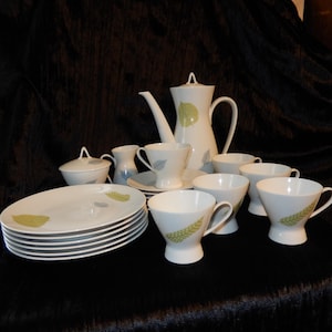 Puede incluir: Juego de té de porcelana blanca con diseño de hojas verdes y grises. El juego incluye una tetera, tazas, platillos, platos, azucarero y jarrita para la leche. El juego de té está dispuesto sobre una superficie oscura.