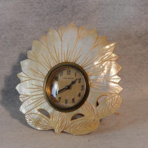 May include: A vintage New Haven clock with a mother-of-pearl sunflower design. The clock face is round with black hands and Roman numeral markings.