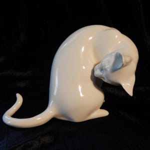 May include: A smooth, glossy, off-white ceramic figurine of a cat in a seated position with its head turned downward toward its side. The cat has a long, curved tail extending outward and a rounded back, captured in a reflective, solid finish.