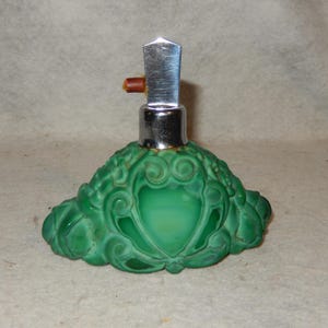 以下が含まれることがあります： シルバーと茶色のストッパーが付いた緑色の香水瓶。ボトルには、装飾的でエンボス加工されたデザインがあります。ボトルのベースは丸みを帯びた形状で、中央にハート型のデザインがあります。ストッパーは長方形です。