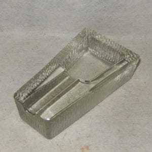 May include: A clear glass ashtray with a rectangular shape and textured surface. The ashtray has a built-in rest for cigarettes and a slightly angled design. The glass has a frosted appearance.
