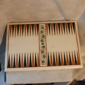 May include: A rectangular backgammon board made of white marble. The board features alternating brown, black, and green triangles along the edges. A central floral design with blue, green, and white accents decorates the center.