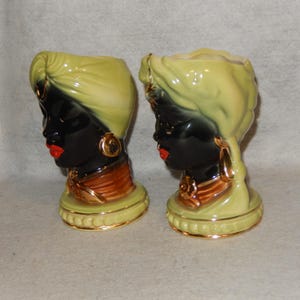 Könnte beinhalten: Zwei Keramikkopfvasen, die jeweils das Profil einer Frau darstellen. Die Gesichter sind schwarz mit roten Lippen, goldenen Ohrringen und einer goldenen Halskette. Die Kopfbedeckungen sind hellgrün und die Basen hellgrün mit goldenen Verzierungen. Ein dekorativer Vintage-Artikel.