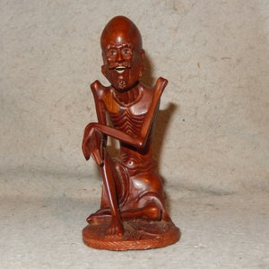 May include: A wooden sculpture of a seated figure with a large head and exaggerated features. The figure is carved in a reddish-brown wood and has a detailed, skeletal appearance.