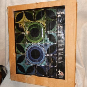 May include: String art kit with a geometric design. The artwork is 16 x 18 inches and features a pattern of circles and squares in shades of green, blue, and teal on a black background. The kit includes a cork board, nails, and threads.