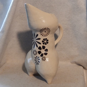 Può includere: Brocca in ceramica bianca con una forma unica e astratta e un manico. La brocca è decorata con motivi floreali stilizzati in nero e blu.