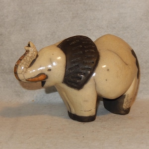 May include: A ceramic elephant figurine with a cream-colored body and dark brown accents. The elephant has a textured, patterned back and a raised trunk. The figurine has a crackle glaze finish, giving it a vintage appearance.