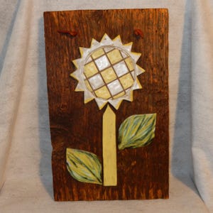 May include: A ceramic sunflower with yellow and white petals and a yellow stem is mounted on a dark brown wooden board.