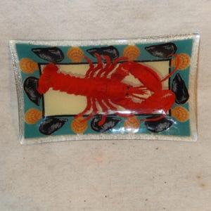 May include: A rectangular glass tray featuring a central illustration of a bright red lobster on a cream background. The border is teal and decorated with a repeating pattern of black mussels and orange seashells, finished with a clear glass edge.