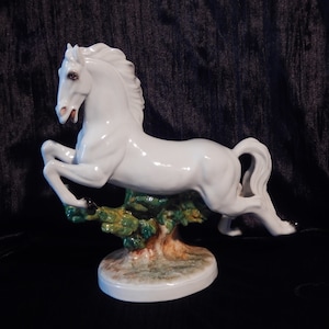 Op de afbeelding: Witte porseleinen figuur van een paard dat over een groene struik springt. Het paard heeft een witte manen en staart en is geschilderd met bruine ogen. De basis van de figuur is geschilderd met groene en bruine details.