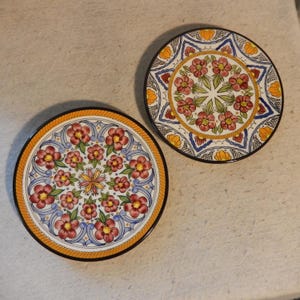 Puede incluir: Dos posavasos decorativos de cerámica con diseños florales. Cada posavasos presenta un patrón floral central en rojo y amarillo, rodeado de elementos decorativos azules y blancos. Los posavasos tienen un borde negro y miden aproximadamente 10 cm de diámetro.