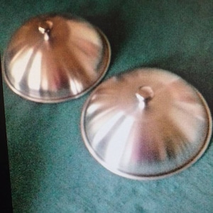 May include: Two silver metal lids for a pot or pan. The lids are round and have a slightly domed shape. They are both shiny and reflect light.