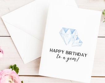May include: White birthday card with a watercolor illustration of two blue diamonds. The card reads "HAPPY BIRTHDAY to a gem!" and is accompanied by a white envelope. Pink flowers are in the background.