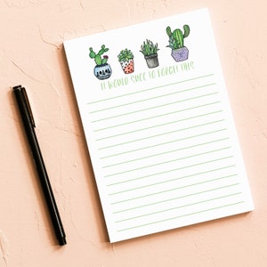 Op de afbeelding: Witte notitieblok met groen gelijnde pagina's en een zwarte pen op een roze ondergrond. Het notitieblok bevat aquarel illustraties van cactussen en vetplanten, met de tekst "IT WOULD SUCC TO FORGET THIS" in het groen.