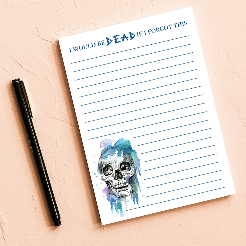 Op de afbeelding: Wit gelijnd notitieblok met de tekst "I WOULD BE DEAD IF I FORGOT THIS" in blauw. Een aquarel schedel illustratie met blauwe en paarse accenten staat onderaan. Een zwarte pen ligt ernaast.