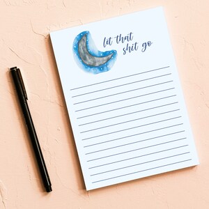 May include: A lined notepad with the phrase "let that shit go" and a watercolor crescent moon design. A black pen rests on a light pink surface next to the notepad.