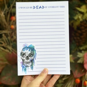 Op de afbeelding: Een gelijnd notitieblok met de tekst "I WOULD BE DEAD IF I FORGOT THIS". Voorzien van een aquarel schedelontwerp met blauwe en groene accenten. Het notitieblok wordt in een hand gehouden.