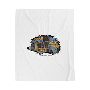 May include: A white blanket featuring a cartoon hedgehog design. The hedgehog is composed of patchwork in shades of yellow, blue, and brown, with the words "SOFT AND SNUGLY" printed below the image.