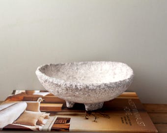 May include: A textured, off-white decorative bowl with three small feet. The bowl is oval-shaped and sits on a book with a soft minimalist theme. The bowl's surface has a rough, handmade appearance, suggesting a natural material.