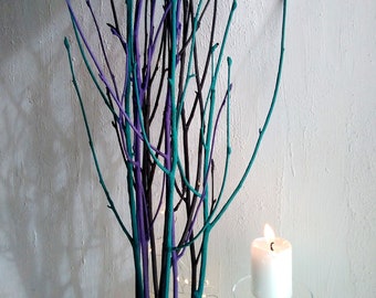 May include: A decorative centerpiece featuring a cluster of painted branches in teal and purple hues. A white candle sits on a clear glass base in front of the branches.