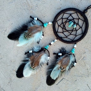 Op de afbeelding: Een zwarte droomvanger met turquoise kralen en veren. De droomvanger heeft een web-achtig ontwerp en hangt aan een touwtje. De veren zijn wit en bruin met turquoise accenten.