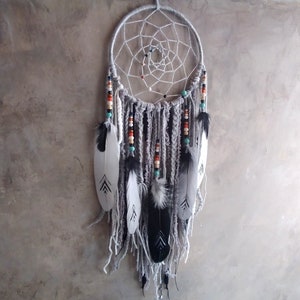 Puede incluir: Un atrapasueños gris y blanco con plumas y cuentas que cuelgan de la parte inferior. El atrapasueños está hecho con una red de macramé blanca y tiene un anillo de madera.