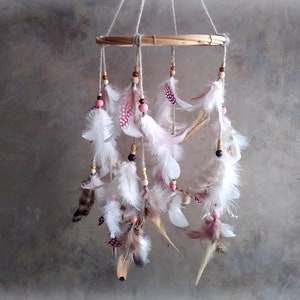 May include: A white and pink feather mobile with wooden beads hanging from a bamboo hoop. The mobile is suspended from the ceiling.