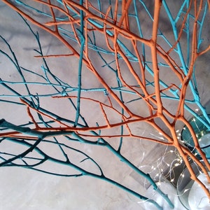 Könnte beinhalten: Eine Gruppe bemalter Zweige in den Farben Türkis und Orange sind in Glasvasen angeordnet. Die Zweige sind miteinander verwoben und schaffen ein einzigartiges und modernes Design. Die Vasen sind mit weißem Sand und kleinen weißen Lichtern gefüllt.