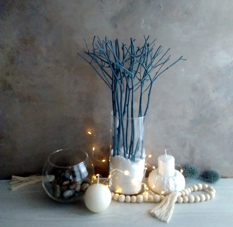 Peut inclure: Un vase en verre rempli de branches peintes en bleu et de sable blanc. Le vase est entour&eacute; de bougies blanches, d'une guirlande de lumi&egrave;res blanches et d'une guirlande de perles en bois.