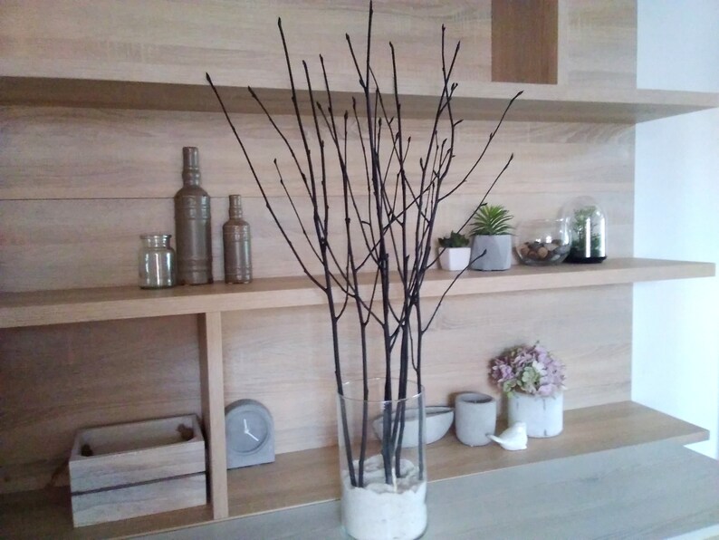 May include: A wooden shelf unit with three shelves, featuring a vase with black branches, two glass bottles, a glass jar, a small wooden box, a clock, and various potted plants.