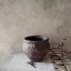 May include: A dark brown, textured ceramic pot with a rough, uneven surface. The pot is sitting on a white surface with a book underneath.