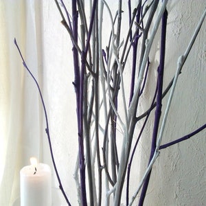 May include: A bunch of twigs painted white and purple, arranged in a vase. A white candle is in the foreground.