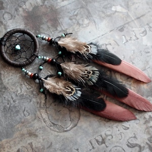 May include: A dreamcatcher ornament featuring a dark brown circular frame with a woven web and a central turquoise bead. Three dangling strands are adorned with small turquoise, black, and red beads, each ending in a long, layered feather.