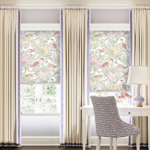 Puede incluir: Dos persianas romanas blancas para ventanas con un patrón floral con pájaros rosas y amarillos. Las persianas están en una habitación con paredes blancas, molduras blancas y cortinas beige. Un escritorio blanco con una silla blanca está en primer plano.