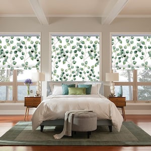 Op de afbeelding: Een slaapkamer met drie ramen met witte rolgordijnen met een groen bladerenpatroon. Het bed heeft witte beddengoed en groene en blauwe kussens. Aan elke kant van het bed staat een houten nachtkastje met een lamp op elk nachtkastje. Er ligt een groen tapijt op de vloer.