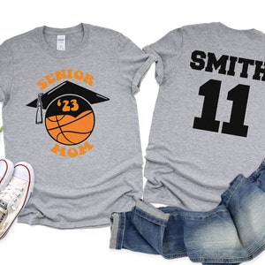 K&ouml;nnte beinhalten: Graues T-Shirt mit dem Text "Senior '23 Mom" und einem Basketball mit einer Abschluss-Kappe darauf. Ein zweites graues T-Shirt mit dem Text "Smith 11" auf der R&uuml;ckseite.
