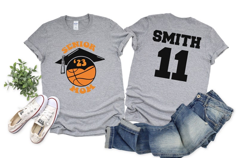 K&ouml;nnte beinhalten: Graues T-Shirt mit dem Text "Senior '23 Mom" und einem Basketball mit einer Abschluss-Kappe darauf. Ein zweites graues T-Shirt mit dem Text "Smith 11" auf der R&uuml;ckseite.