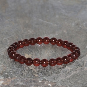 Puede incluir: Un brazalete de piedras preciosas rojas hecho de cuentas redondas. El brazalete está sobre un fondo gris.