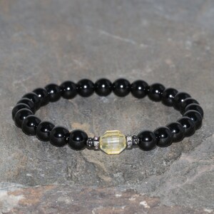 Puede incluir: Pulsera de cuentas negras con una gema amarilla facetada en el centro. La pulsera presenta pequeñas cuentas negras redondas y brillantes y un acento plateado decorativo a cada lado de la piedra amarilla. La pulsera se muestra sobre una superficie de piedra gris.