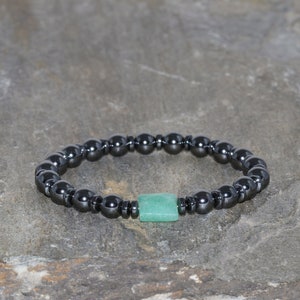Könnte beinhalten: Ein Armband aus schwarzen Hämatitperlen mit einem quadratischen grünen Stein als Anhänger.